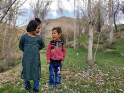 Two young children interact joyfully in a natural setting surrounded by bare trees and patches of grass. The older child wears a teal patterned dress, while the younger child dons a red and green jacket with blue pants. Both children have bright expressions, suggesting a playful conversation.