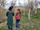 Two young children interact joyfully in a natural setting surrounded by bare trees and patches of grass. The older child wears a teal patterned dress, while the younger child dons a red and green jacket with blue pants. Both children have bright expressions, suggesting a playful conversation.