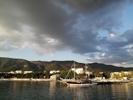 A coastline with a large, dark wooden sailing ship docked near a sandy shore. In the background, there are forested hills and a small town with several buildings nestled among the trees. The sky is filled with dramatic, dark clouds and patches of blue.