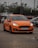 A bright orange car is parked in an outdoor lot with a few other vehicles nearby. The setting is somewhat cloudy, and the car has its headlights on, highlighting its sleek design. Trees and a dull sky form the backdrop, enhancing the focus on the car.