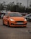 A bright orange car is parked in an outdoor lot with a few other vehicles nearby. The setting is somewhat cloudy, and the car has its headlights on, highlighting its sleek design. Trees and a dull sky form the backdrop, enhancing the focus on the car.
