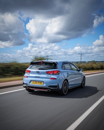 A light blue hatchback car is driving on a highway under a partly cloudy sky. The car features sleek, sporty design elements and a visible rear spoiler. The surrounding landscape includes green bushes lining the road.