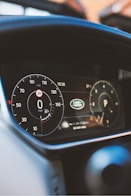 A close-up view of a car dashboard showing the speedometer, odometer, and fuel gauge. The speedometer reads 0 mph, and the odometer displays 24449 miles. Logos and electronic displays are visible, indicating the car's status and brand.