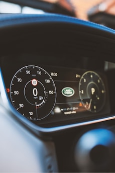 A close-up view of a car dashboard showing the speedometer, odometer, and fuel gauge. The speedometer reads 0 mph, and the odometer displays 24449 miles. Logos and electronic displays are visible, indicating the car's status and brand.