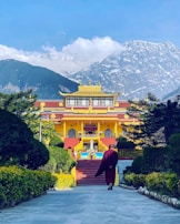 A person in traditional clothing walks towards a vibrant yellow and red Buddhist monastery surrounded by green foliage. Snow-capped mountains tower in the background under a clear blue sky.