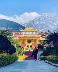 A person in traditional clothing walks towards a vibrant yellow and red Buddhist monastery surrounded by green foliage. Snow-capped mountains tower in the background under a clear blue sky.