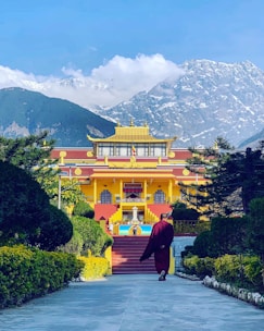 A person in traditional clothing walks towards a vibrant yellow and red Buddhist monastery surrounded by green foliage. Snow-capped mountains tower in the background under a clear blue sky.