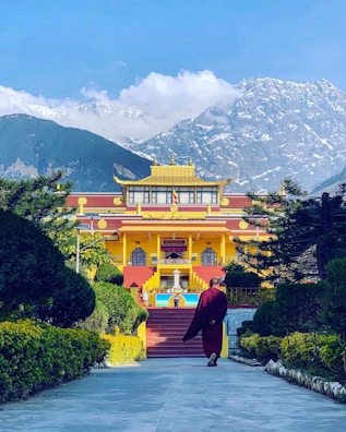 A person in traditional clothing walks towards a vibrant yellow and red Buddhist monastery surrounded by green foliage. Snow-capped mountains tower in the background under a clear blue sky.