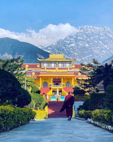 A person in traditional clothing walks towards a vibrant yellow and red Buddhist monastery surrounded by green foliage. Snow-capped mountains tower in the background under a clear blue sky.