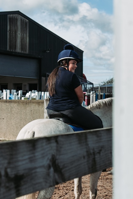 A person wearing a blue riding helmet and a dark shirt is sitting on a white horse. The setting appears to be an outdoor paddock with a wooden fence in the foreground and a large barn or stable in the background. The sky is partly cloudy, and there is an assortment of riding equipment along the fence.