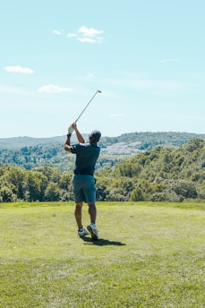 A person is performing a golf swing on a lush green golf course under a clear blue sky. Rolling hills with dense trees form the backdrop, creating a serene and expansive view.