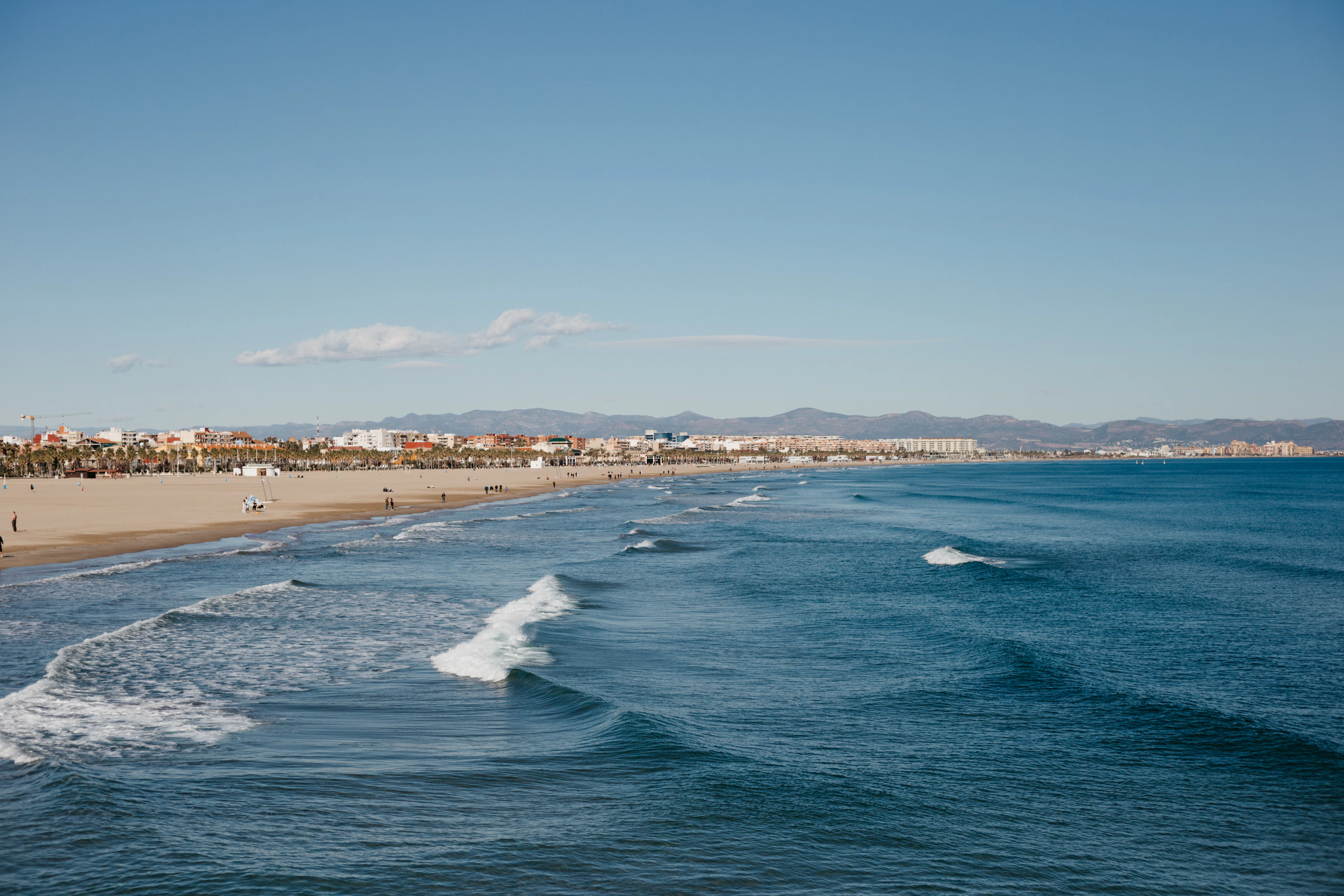 Waves roll onto Valencia's long sandy beach, the city skyline and hills visible in the distance.