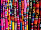 A collection of vibrant, colorful textile fabrics arranged vertically. The fabrics feature intricate patterns and designs, including geometric shapes and various traditional motifs. The array of colors includes bright pinks, oranges, blues, greens, and purples.