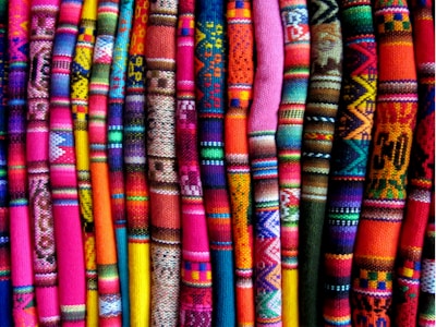 A collection of vibrant, colorful textile fabrics arranged vertically. The fabrics feature intricate patterns and designs, including geometric shapes and various traditional motifs. The array of colors includes bright pinks, oranges, blues, greens, and purples.