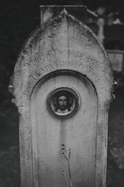 An old and weathered tombstone with Cyrillic inscriptions is prominently displayed. In the center of the tombstone, there is a small framed circular image depicting a religious figure, likely Jesus Christ. The stone appears aged, with stains and cracks, and is surrounded by a dark, blurred background that hints at more tombstones and possible foliage.