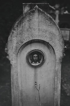 An old and weathered tombstone with Cyrillic inscriptions is prominently displayed. In the center of the tombstone, there is a small framed circular image depicting a religious figure, likely Jesus Christ. The stone appears aged, with stains and cracks, and is surrounded by a dark, blurred background that hints at more tombstones and possible foliage.