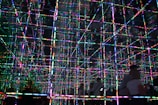 A mesmerizing grid of multicolored LED lights creates a three-dimensional effect with reflections across multiple mirrored surfaces. The lines of light intersect at right angles, forming a complex pattern that appears infinite and immersive. Two figures are visible in the foreground, partially silhouetted against the vibrant display.