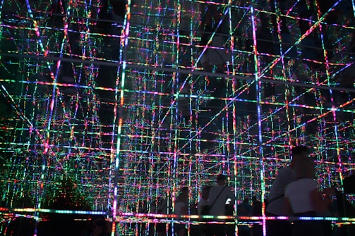 A mesmerizing grid of multicolored LED lights creates a three-dimensional effect with reflections across multiple mirrored surfaces. The lines of light intersect at right angles, forming a complex pattern that appears infinite and immersive. Two figures are visible in the foreground, partially silhouetted against the vibrant display.