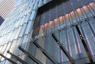 A modern skyscraper facade with reflective glass panels, featuring large lettering that spells 'ONE WORLD.' The building design includes vertical lines and a sleek, metallic structure. Lights are positioned between glass sections, and the facade captures reflections of the sky and other buildings.