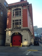 A red-brick building with a prominent firehouse entrance and an American flag hanging above the door. The facade is adorned with simple architectural details, and there is a golden emblem above the entrance. The surrounding area includes urban buildings with visible city streets and vehicles parked nearby.