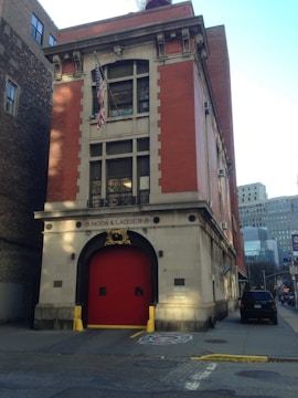 A red-brick building with a prominent firehouse entrance and an American flag hanging above the door. The facade is adorned with simple architectural details, and there is a golden emblem above the entrance. The surrounding area includes urban buildings with visible city streets and vehicles parked nearby.