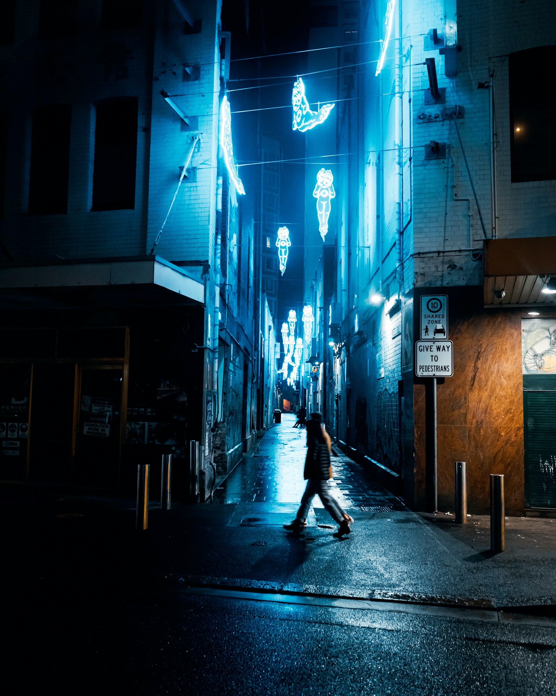 A quiet alley bathed in moody blue and purple neon lights, steam rising from a small street vendor's cart under the rain.