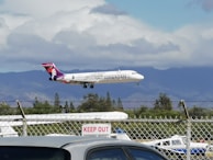 An airplane with Hawaiian Airlines branding is captured mid-flight against a backdrop of mountains and partial cloud cover. The scene includes a chain-link fence with a 'Keep Out' sign in the foreground. The sky is primarily cloudy with hints of blue throughout.