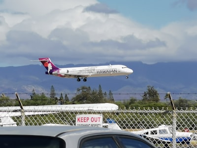 An airplane with Hawaiian Airlines branding is captured mid-flight against a backdrop of mountains and partial cloud cover. The scene includes a chain-link fence with a 'Keep Out' sign in the foreground. The sky is primarily cloudy with hints of blue throughout.