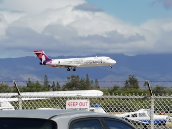 An airplane with Hawaiian Airlines branding is captured mid-flight against a backdrop of mountains and partial cloud cover. The scene includes a chain-link fence with a 'Keep Out' sign in the foreground. The sky is primarily cloudy with hints of blue throughout.
