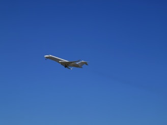 A sleek, white private jet is captured in mid-flight against a clear, vibrant blue sky. The aircraft's wings are extended, and its landing gear is visible.
