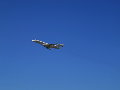 A sleek, white private jet is captured in mid-flight against a clear, vibrant blue sky. The aircraft's wings are extended, and its landing gear is visible.