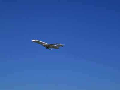 A sleek, white private jet is captured in mid-flight against a clear, vibrant blue sky. The aircraft's wings are extended, and its landing gear is visible.