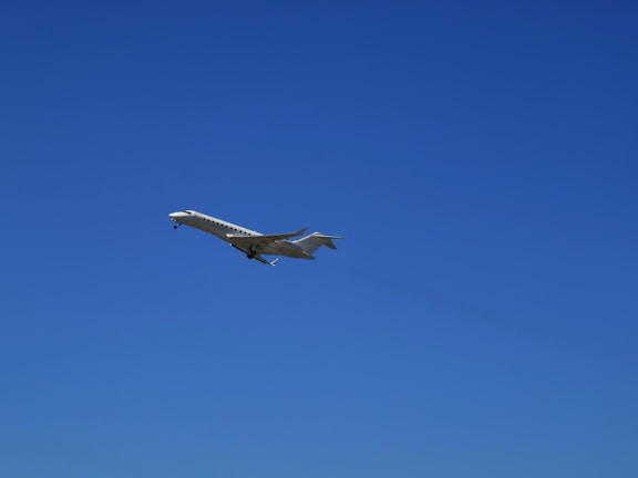 A sleek, white private jet is captured in mid-flight against a clear, vibrant blue sky. The aircraft's wings are extended, and its landing gear is visible.