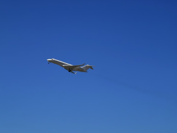 A sleek, white private jet is captured in mid-flight against a clear, vibrant blue sky. The aircraft's wings are extended, and its landing gear is visible.