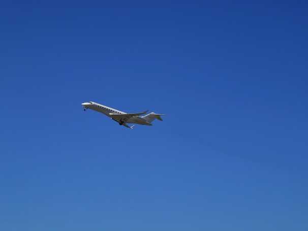 A sleek, white private jet is captured in mid-flight against a clear, vibrant blue sky. The aircraft's wings are extended, and its landing gear is visible.