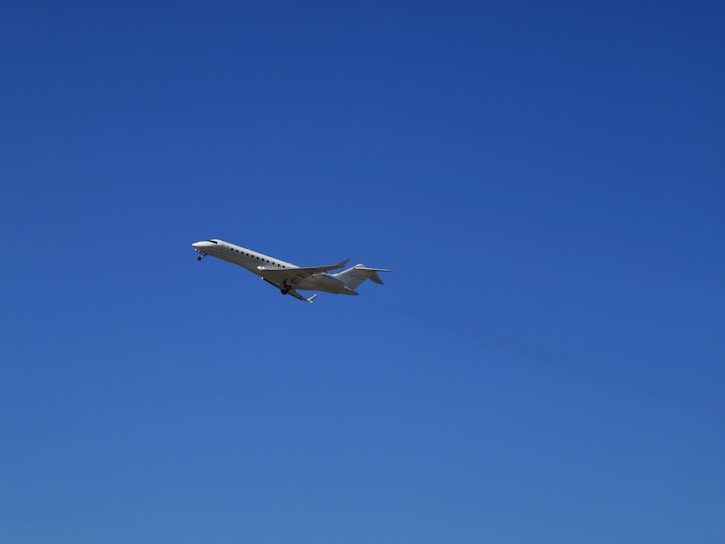 A sleek, white private jet is captured in mid-flight against a clear, vibrant blue sky. The aircraft's wings are extended, and its landing gear is visible.