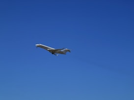 A sleek, white private jet is captured in mid-flight against a clear, vibrant blue sky. The aircraft's wings are extended, and its landing gear is visible.