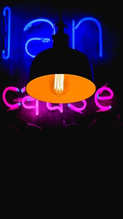 A dark setting illuminated by a hanging lamp with an exposed bulb, casting a warm amber glow. Behind the lamp, neon signs are visible, with blue and pink letters glowing against the dark backdrop.
