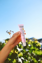 A hand holding a pink tube of sunscreen labeled 'Everyday Humans Rosé S'il Vous Plait SPF30 Gentle Mineral Sunscreen'. The background features lush green leaves and a clear blue sky, conveying a bright and natural setting.