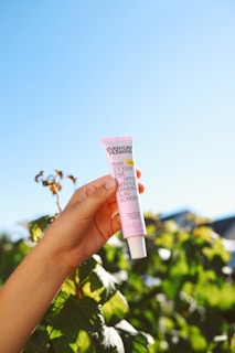 A hand holding a pink tube of sunscreen labeled 'Everyday Humans Rosé S'il Vous Plait SPF30 Gentle Mineral Sunscreen'. The background features lush green leaves and a clear blue sky, conveying a bright and natural setting.