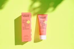 Two Everyday Humans SPF50 Body Sunscreen products are displayed against a bright green background. The product on the left is in its packaging box, and the one on the right is the tube. Both items are in a coral color, and shadows are cast on the background, suggesting direct light.