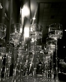 A collection of tall, elegant wine glasses with long stems arranged closely together, creating a sense of opulence and luxury. The image captures reflections and refractions of light through the transparent glass, adding depth to the scene. In the background, a softly-lit room can be seen, contributing to a sophisticated and serene atmosphere.