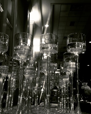 A collection of tall, elegant wine glasses with long stems arranged closely together, creating a sense of opulence and luxury. The image captures reflections and refractions of light through the transparent glass, adding depth to the scene. In the background, a softly-lit room can be seen, contributing to a sophisticated and serene atmosphere.