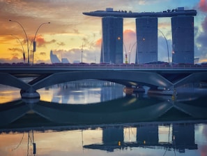 A modern cityscape featuring a prominent luxury hotel complex with three towering structures topped by a unique, boat-shaped sky park. In the foreground, a bridge extends across a calm body of water that reflects the vibrant colors of the sunset and the buildings. The sky is a mix of soft orange, pink, and blue hues, adding a serene atmosphere to the scene.