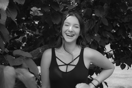 A person with long hair and a joyful expression, standing against a backdrop of dense foliage. The monochrome image highlights the contrast between the person's light skin and dark clothing.