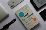 A flat lay arrangement featuring a book titled 'Lost Connections' by Johann Hari surrounded by various electronic devices on a white surface. To the top left is a silver laptop partially visible. Next to it, there is an iPhone with the Apple logo and a dual camera visible on its back. A black smartwatch with a strap is placed below the laptop. To the right, there is an Amazon Kindle displaying text and images on its screen. Near the Kindle, a black device, possibly another phone, lies with part of a case visible.