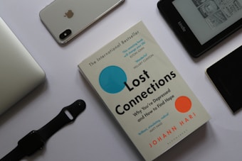A flat lay arrangement featuring a book titled 'Lost Connections' by Johann Hari surrounded by various electronic devices on a white surface. To the top left is a silver laptop partially visible. Next to it, there is an iPhone with the Apple logo and a dual camera visible on its back. A black smartwatch with a strap is placed below the laptop. To the right, there is an Amazon Kindle displaying text and images on its screen. Near the Kindle, a black device, possibly another phone, lies with part of a case visible.