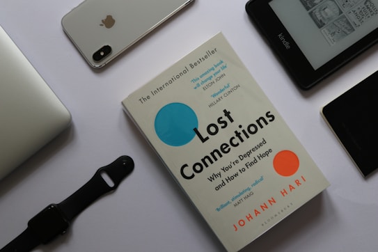 A flat lay arrangement featuring a book titled 'Lost Connections' by Johann Hari surrounded by various electronic devices on a white surface. To the top left is a silver laptop partially visible. Next to it, there is an iPhone with the Apple logo and a dual camera visible on its back. A black smartwatch with a strap is placed below the laptop. To the right, there is an Amazon Kindle displaying text and images on its screen. Near the Kindle, a black device, possibly another phone, lies with part of a case visible.