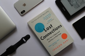 A flat lay arrangement featuring a book titled 'Lost Connections' by Johann Hari surrounded by various electronic devices on a white surface. To the top left is a silver laptop partially visible. Next to it, there is an iPhone with the Apple logo and a dual camera visible on its back. A black smartwatch with a strap is placed below the laptop. To the right, there is an Amazon Kindle displaying text and images on its screen. Near the Kindle, a black device, possibly another phone, lies with part of a case visible.