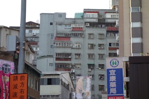 A tall, rectangular residential building with multiple air conditioning units and balconies, surrounded by smaller commercial buildings and signs with Chinese characters. The structure shows signs of wear, and laundry can be seen hanging on several balconies.